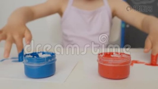 一个小可爱的女孩坐在桌子旁，用明亮的手指颜料画纸，把她的手指浸在颜料罐里。视频