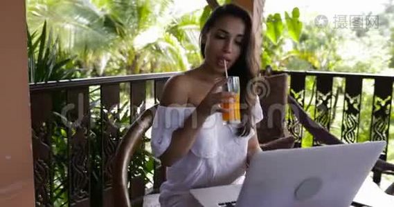 女孩用笔记本电脑坐在夏天的露台上，喝橙汁吃早餐，年轻女人聊天。视频