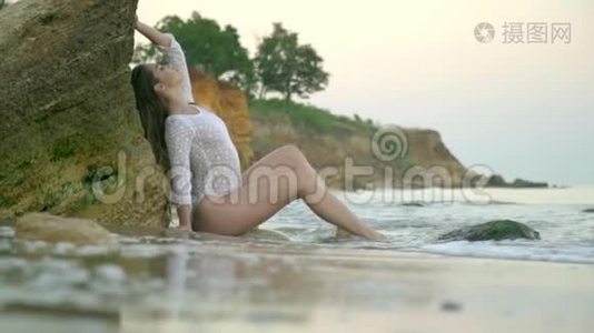 神秘的苗条女孩穿着白色连衣裙，坐在海角中部的一块岩石下，户外视频