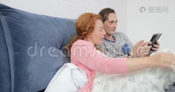 使用数码平板电脑躺在床上的情侣，男人和女人在卧室里聊天浏览互联网视频