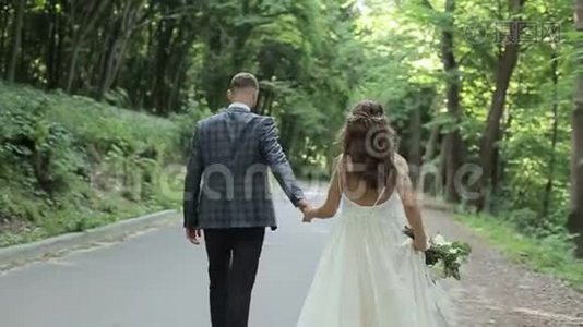 一对漂亮的已婚夫妇走在公园里牵着他们的手。 结婚礼服。 背景上的绿树视频