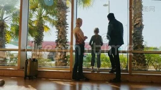 机场候机楼的年轻家庭。 站在窗口，看飞机。 外面，一个阳光明媚的日子，棕榈树生长视频
