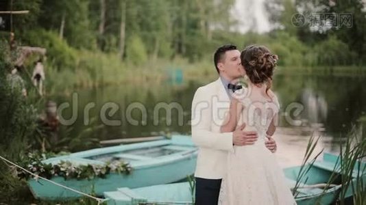 美丽的恋人在一个美丽的地方亲吻一个池塘，池塘上有蓝色的船。 新娘和新郎享受他们的婚礼视频