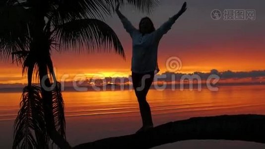 一个女孩站在海滩上红日落的背景下的棕榈树上视频