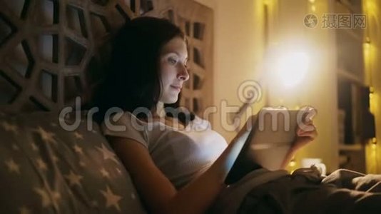 年轻的笑脸女人，晚上躺在家里的床上，用平板电脑上网购物视频