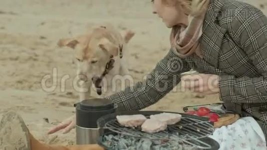 穿着外套的年轻女子坐在河边、海边的海滩上，野餐，在烤架上煮蔬菜和肉视频