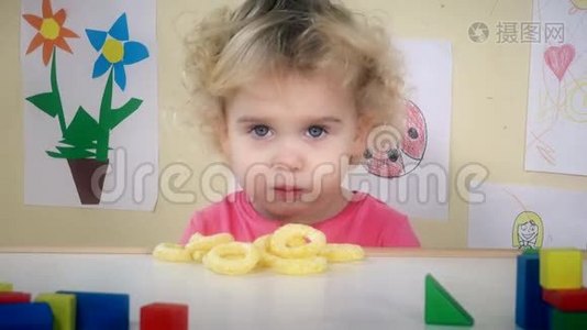 小女孩吃玉米圈，坐在她的桌子旁边，玩具和图画视频