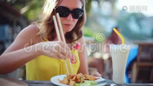 年轻优雅的幸福女人戴着太阳镜吃垫泰国炒面加鸡蛋，泰国菜。 坐在咖啡馆里视频