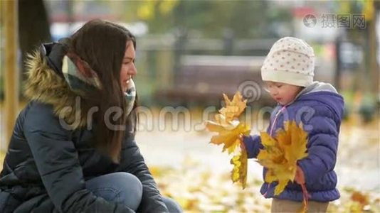 快乐的年轻妈妈和她的小女儿在秋天的公园里玩，妈妈和女孩扔树叶和笑视频