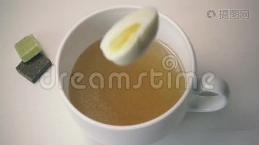 鸡蛋掉进清汤的白色杯子里。 慢动作视频