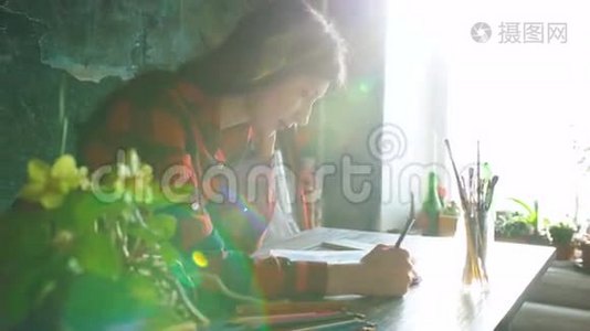 年轻的女艺术家用铅笔在纸笔记本上画。 明亮的太阳从窗户照进来视频