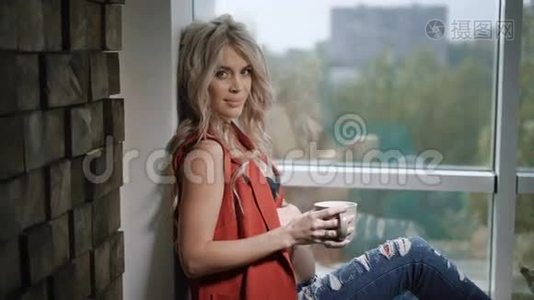 穿着红色衬衫和蓝色牛仔裤的美丽怀孕金发女人坐在靠近窗户和墙壁的窗台上视频