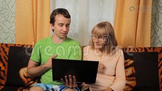 成年男性教为笔记本电脑工作的老妇人。 母子俩坐在家里的沙发上视频