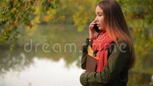 美丽的学生女在湖边的秋天花园里用手机说话，美丽的女孩笑着，公共公园视频