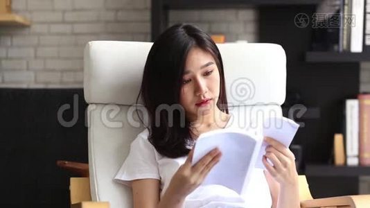 美丽的肖像，年轻的亚洲女人放松坐在客厅看书在家，女孩学习文学。视频
