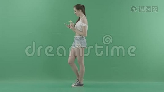 年轻女性在绿色背景下喝鸡尾酒时在智能手机上发短信视频