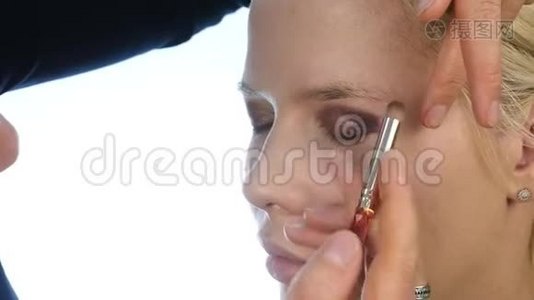 女性造型师用特殊的画笔在金发新娘的眼皮上涂上深色的眼影。 婚礼化妆。 慢动作视频