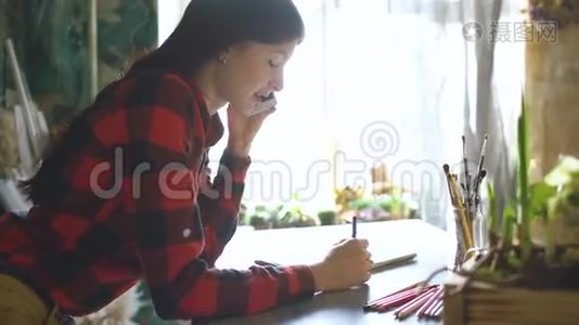 年轻的女艺术家用铅笔在纸笔记本上画。 女孩微笑，打电话视频