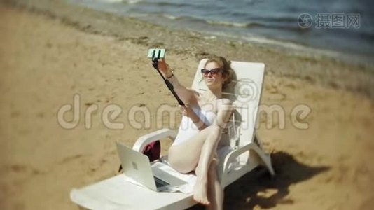 一个穿着白色比基尼的年轻女孩躺在沙滩上的躺椅上晒太阳，还在智能手机上拍照视频