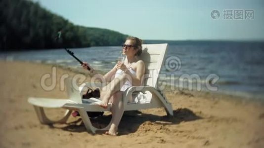 一个穿着白色比基尼的年轻女孩躺在沙滩上的躺椅上晒太阳，还在智能手机上拍照视频