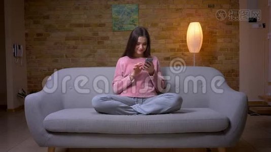 一位年轻漂亮的褐发白种人女性在舒适的沙发上用电话微笑特写镜头视频