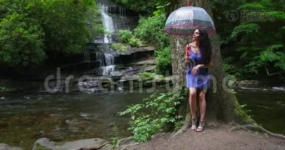 一个年轻的女人拿着雨伞站在瀑布边的森林里，看着落水。视频