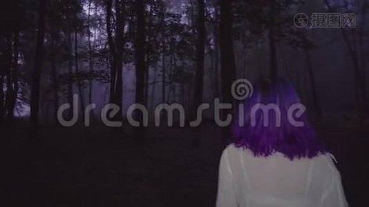 在黑暗森林中漫步的白色衬衫和紫色头发的美丽女人的后景-惊悚的场景。视频