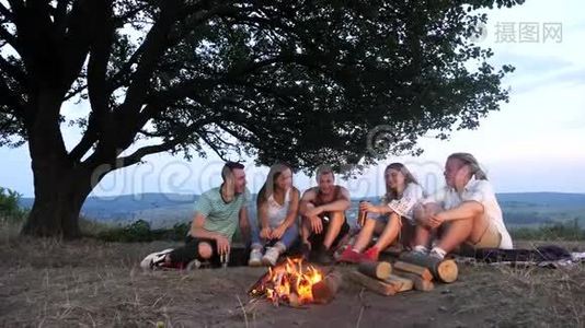 一群朋友坐在篝火旁，喝着热饮料和交谈视频