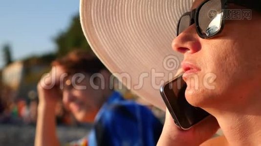 4K女人戴着白色宽边帽子和太阳镜，在海滩上用她的手机，慢动作。视频