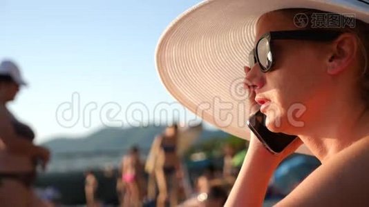 4K女人戴着白色宽边帽子和太阳镜，在海滩上用她的手机，慢动作。视频