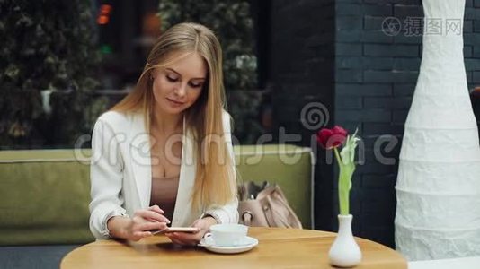 迷人的金发女人在她的智能手机上读到一些东西，坐在咖啡馆的桌子旁。视频