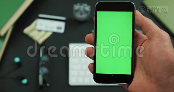男人拿着一个绿色屏幕的黑色智能手机在工作桌上视频
