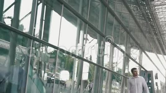 英俊的商人戴着眼镜和手提箱在现代机场航站楼玻璃墙背景。 旅行者视频