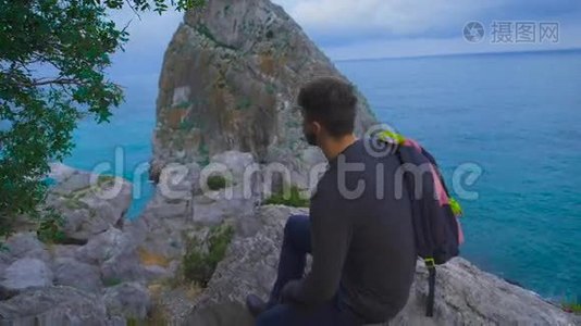 年轻的嬉皮士坐在海边的岩石山上，背上背着背包的男人视频