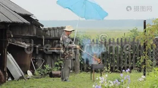 带着雨伞的人在火旁烧烤。 雨在下，一座背景有栅栏的老建筑视频