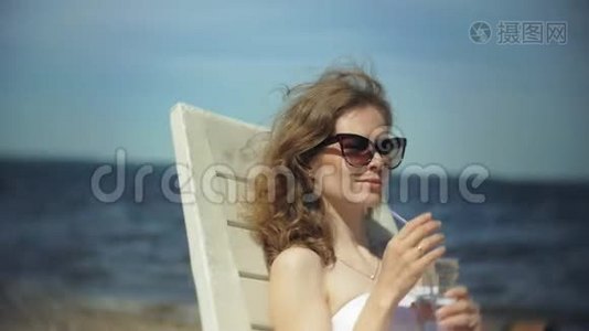 一个穿着白色比基尼的年轻女孩躺在海边沙滩上的休息室里晒太阳，喝着鸡尾酒视频