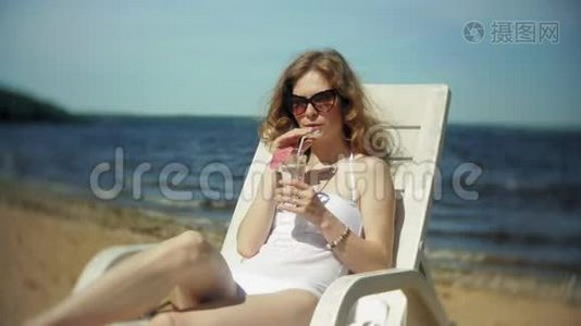 一个穿着白色比基尼的年轻女孩躺在海边沙滩上的休息室里晒太阳，喝着鸡尾酒视频