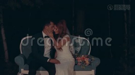 新郎和新娘在晚上拥抱和亲吻，坐在黑暗森林的沙发上。视频