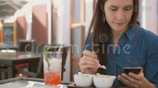 布鲁内特女孩坐在外面的咖啡馆里吃冰淇淋时使用智能手机。 4k，视频