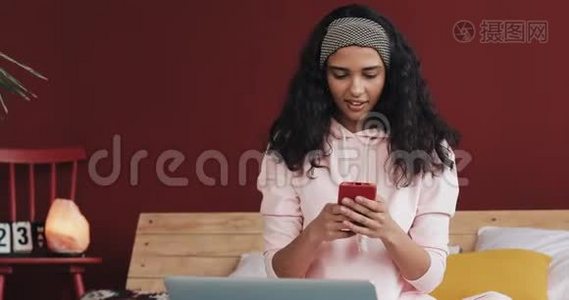 快乐的非裔美国女孩聊天和短信智能手机坐在床上在家里。 年轻女子视频