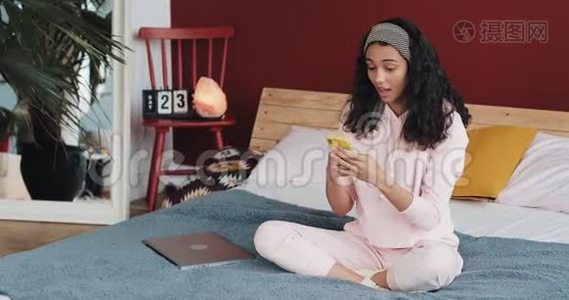 快乐的非裔美国女孩聊天和短信智能手机坐在床上在家里。 年轻女子视频
