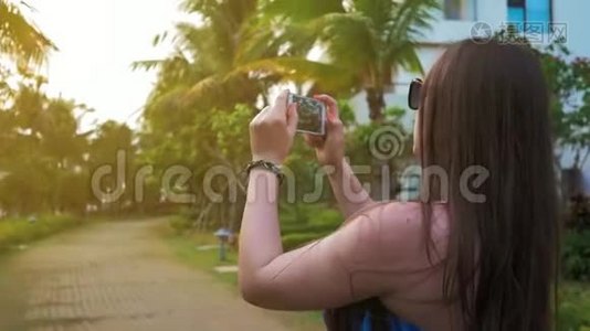 漂亮的快乐女孩戴着眼镜和一件蓝色的连衣裙，正在手机上拍摄夕阳的照片。 绿色的手掌和视频