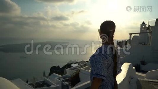 快乐的女人穿着白色和蓝色的裙子在希腊的桑托里尼享受她的假期。 从卡尔德拉和爱琴海观景视频
