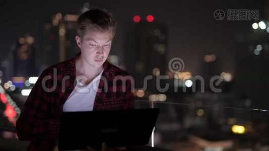 年轻的帅哥晚上用笔记本电脑观看城市景色视频