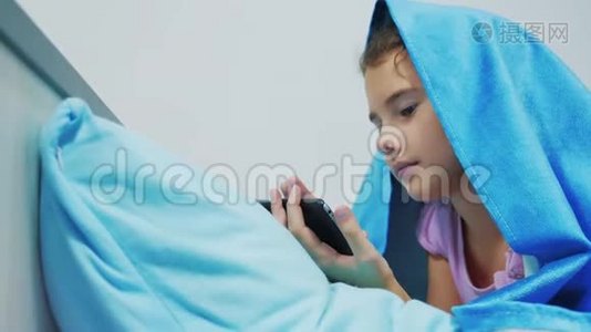 带智能手机的毯子下可爱女孩的肖像。 小女孩在社交媒体毯子下玩网络游戏。 女孩视频