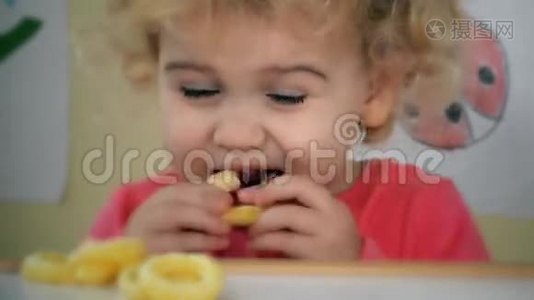小女孩吃玉米圈，坐在桌子旁边，有玩具和图画在墙上视频