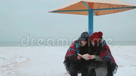 一个女孩和一个男人坐在海滩的天篷下，上面铺着一个格子，在看书。视频