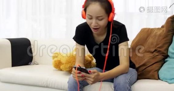 美丽的亚洲女人正在听音乐耳机，在家里的沙发上使用智能手机，放松生活方式视频