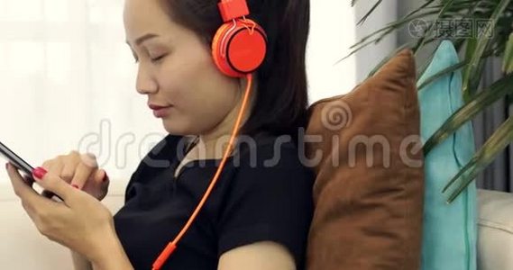 美丽的亚洲女人正在听音乐耳机，在家里的沙发上使用智能手机，放松生活方式视频