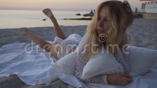 那个女孩躺在沙滩上枕头和毯子之间。 日落，浪漫，慢动作，特写。 美丽的视频